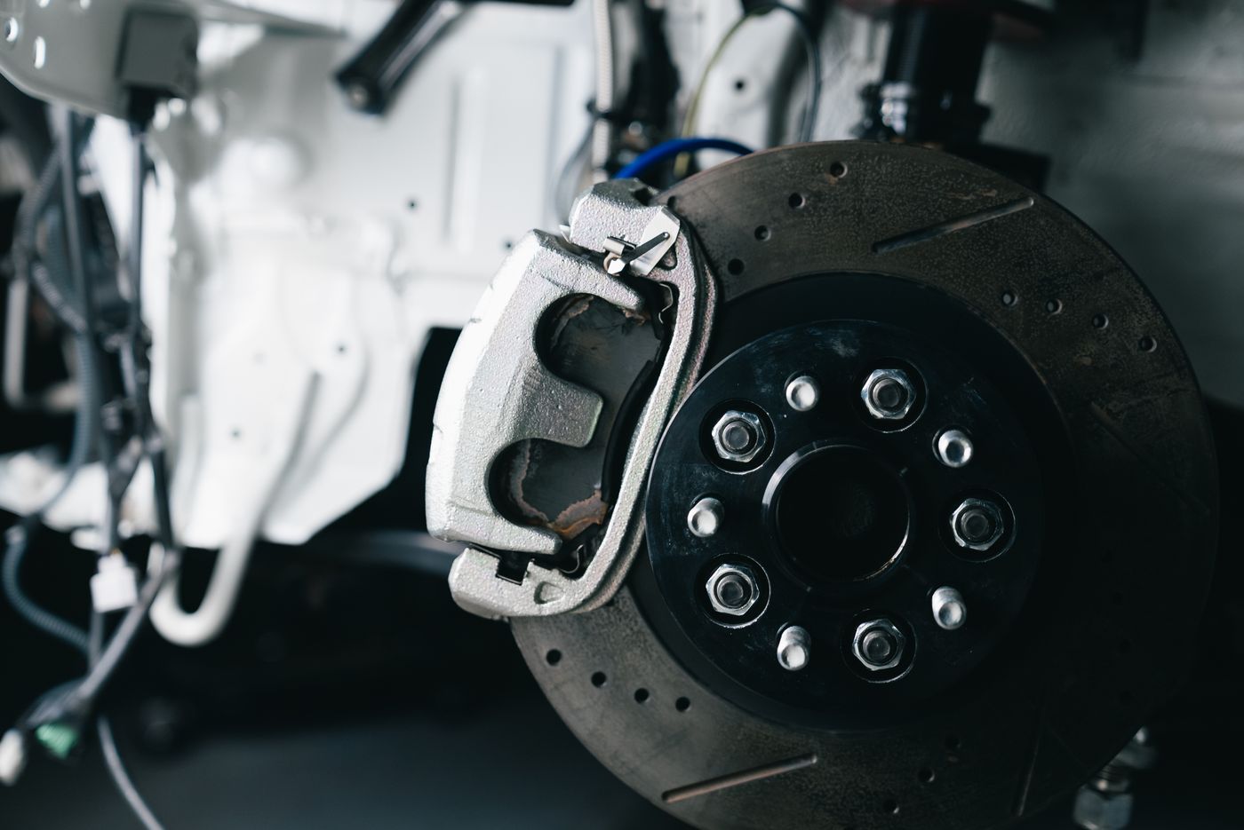 A silver brake caliper over a drilled and slotted disc on a black hub with eight wheel studs and chrome nuts, white bodywork and loose wiring blurred behind.