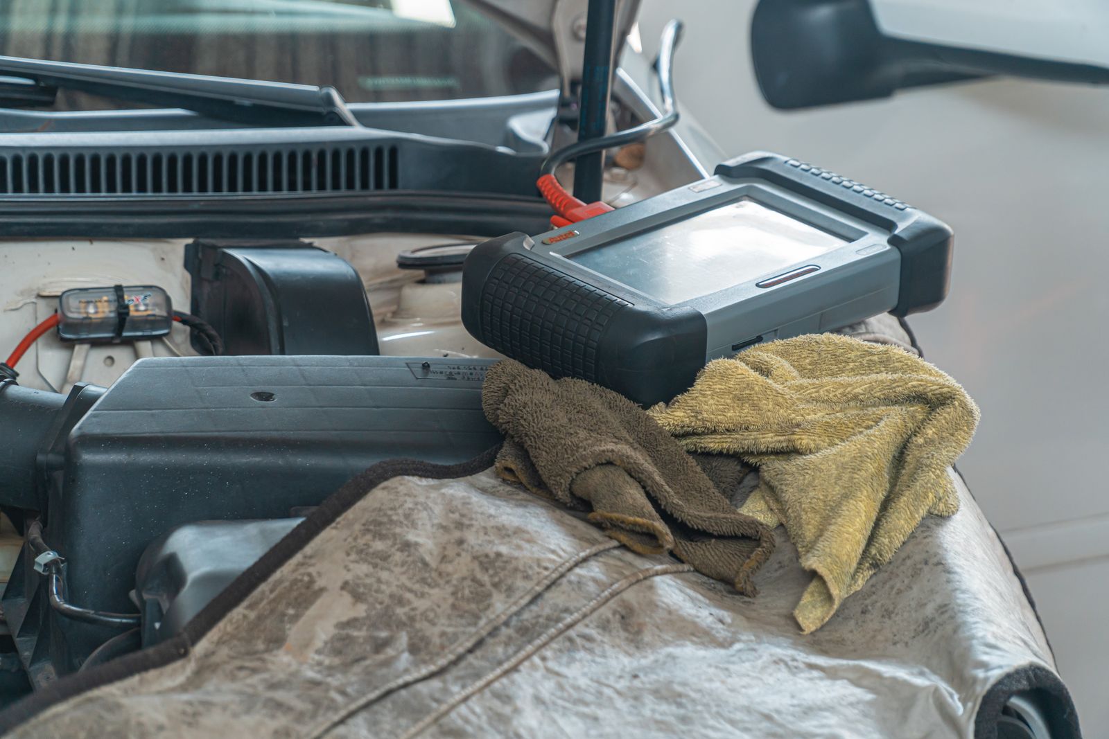 A grey-and-black rugged handheld diagnostic scanner with a red lead resting on towels draped over the open bonnet of a white car.